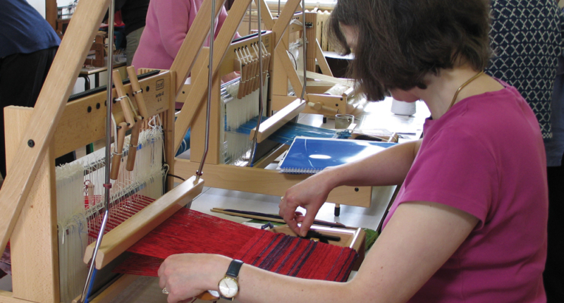 A weaver using a loom in weaving class- others looms and weavers in the background